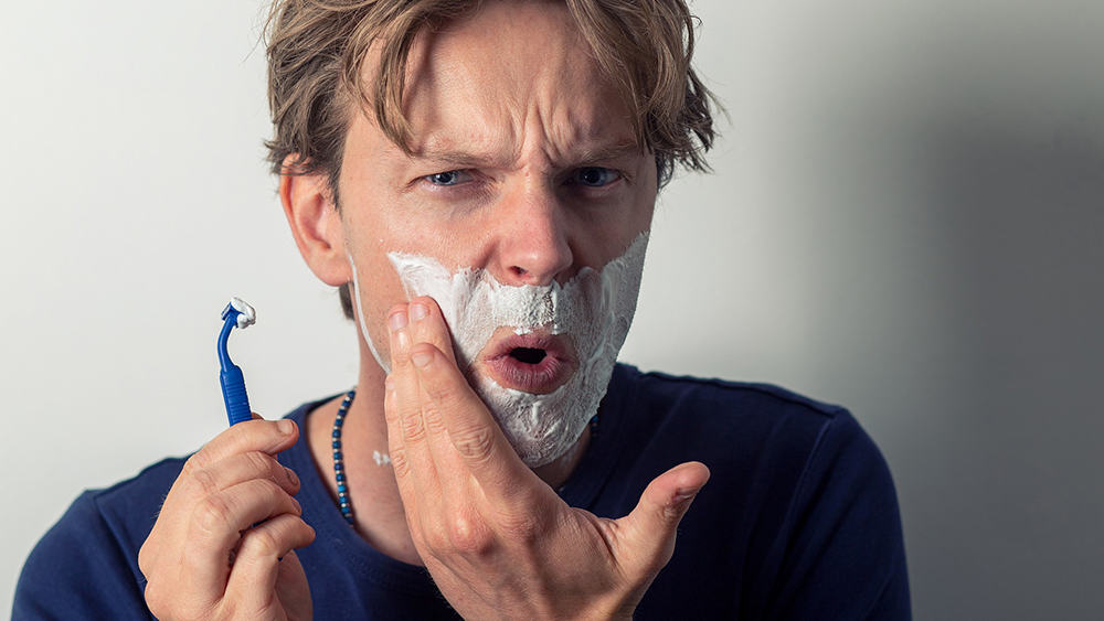 Afbeelding van een man met scheerschuim op zijn gezicht en een scheermesje in zijn hand.