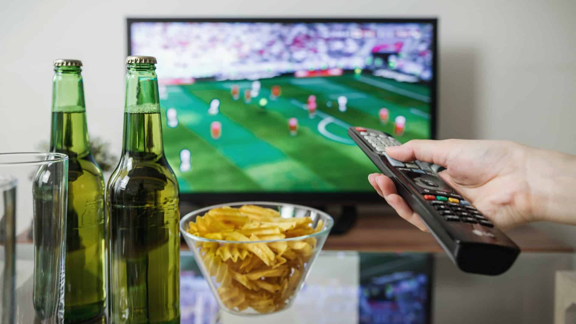 Afbeelding van een televisie op de achtergrond en op het tafeltje staan twee flesjes bier en een bakje chips.  