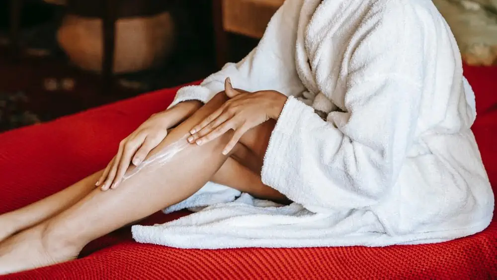 Afbeelding van een persoon in ochtendjas die crème op de benen smeert. 