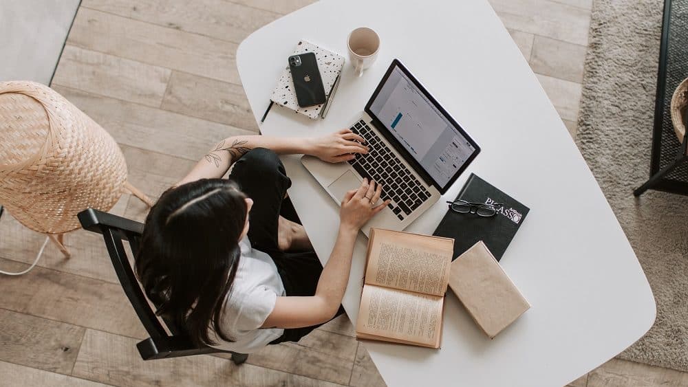 Afbeelding van een persoon die achter haar laptop zit met op haar bureau boeken, een agenda, smartphone en een mok. 
