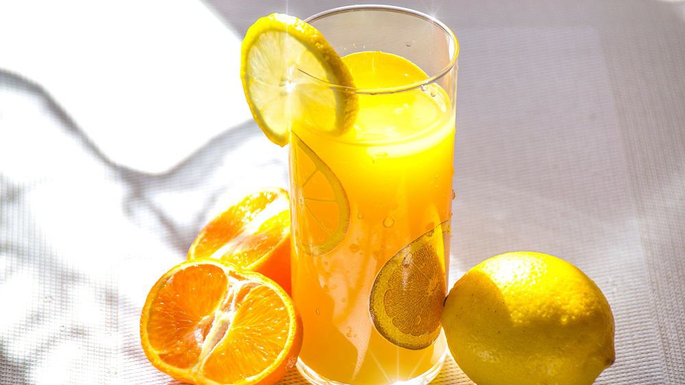 Afbeelding van een glas met sinaasappel-citroensap. Naast het glas ligt een citroen en een sinaasappel in twee stukken. 