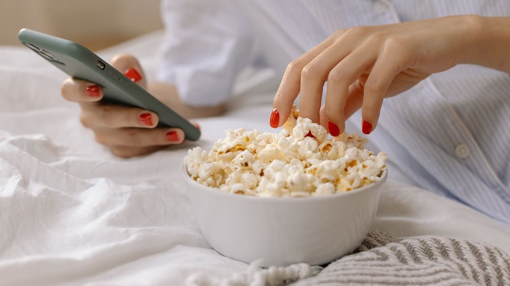 Afbeelding van iemand die met de ene hand een smartphone vastheeft en met de andere hand graait in een bakje popcorn.