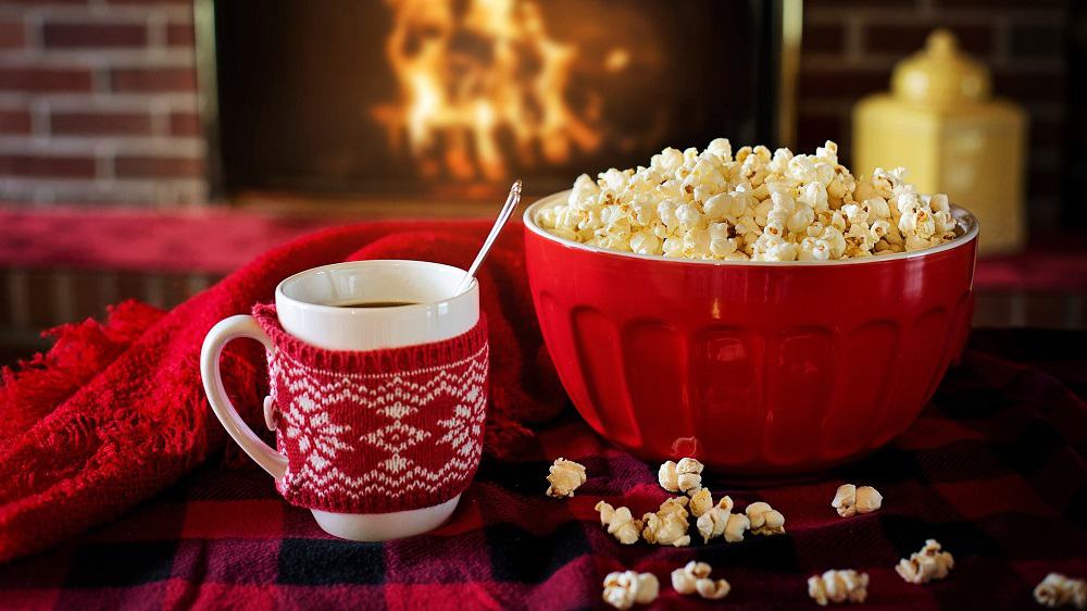 Afbeelding van een grote bak popcorn, een mok met warme chocolademelk en op de achtergrond een open haard.  