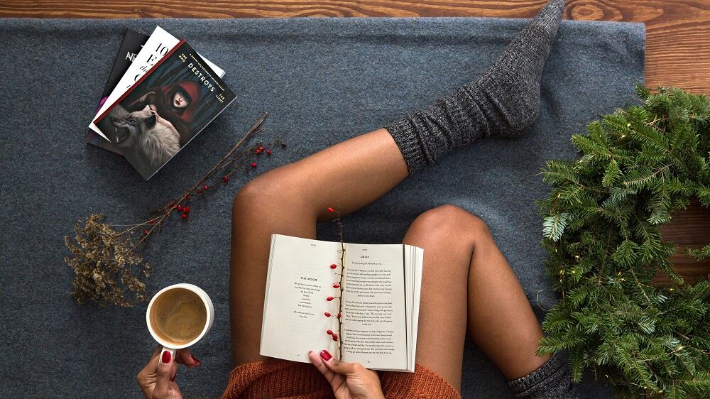 Foto van bovenaf van iemand die op een kleed zit met met dikke sokken aan, een boek op schoot en koffie in de hand.