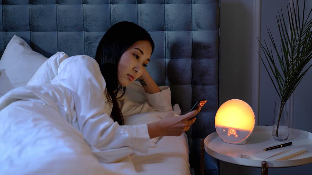 Afbeelding van iemand die in bed zit en op zijn telefoon kijkt. Op het nachtkastje naast het bed staat een verlichte wekker, een plant, een schrift en een pen. 